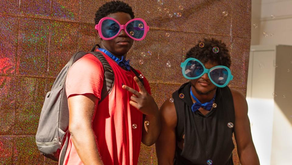 Two students take a photo together while wearing oversized sunglasses in front of a glitter background