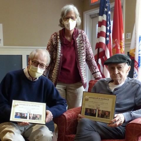 Photo of two veterans sitting in chairs with awards and Susan Gold posing with them